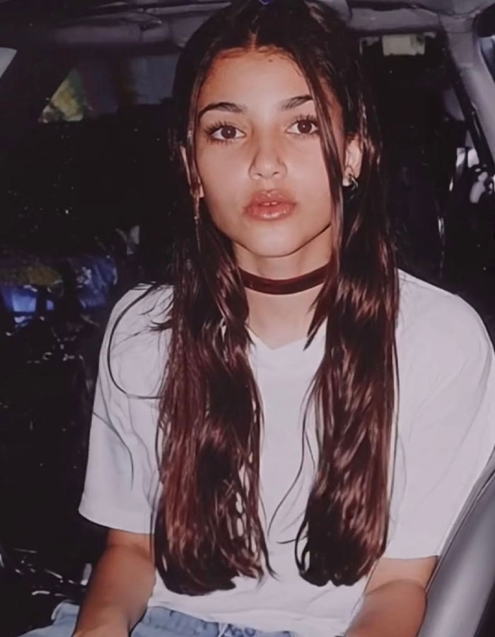 Mujer joven con largo cabello castaño y gargantilla sentada en un automóvil, vinculada al retroceso de Kim Kardashian y la discusión de cirugías de plástico.