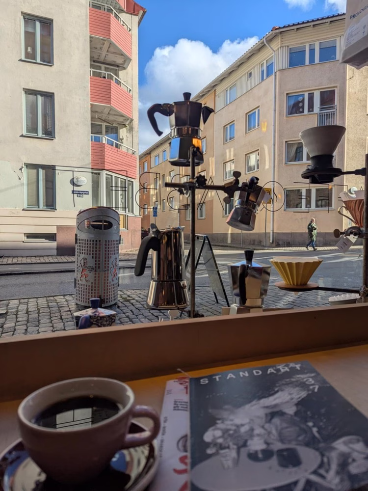Una vista desde una ventana de café en Gotemburgo, Suecia. Fuera de la ventana hay grandes edificios y un cielo azul brillante. Frente a la ventana hay un bar de madera, cubierto con una taza de café y una revista de café titulada