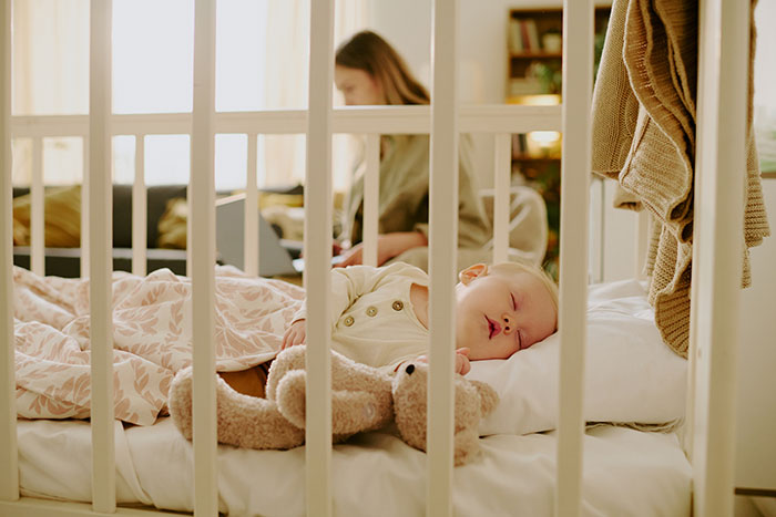 El bebé se queda dormido en cuna con osito de peluche, mientras que el miembro de la familia trabaja en la computadora portátil en el fondo durante el cuidado de la niñera.