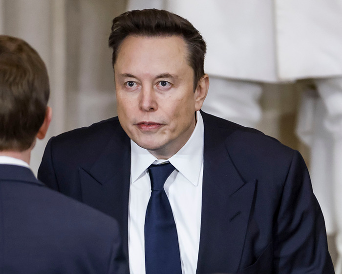 Elon Musk con traje oscuro y corbata se dedicó a una conversación, enfrentando a otra persona en un entorno interior formal.