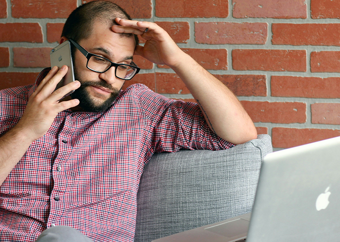 El ingeniero en gafas parece frustrado en una llamada mientras está sentado en un sofá con la computadora portátil, reflexionando sobre un tratamiento difícil.