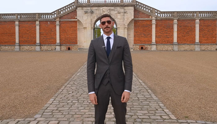 Hombre con traje oscuro y gafas de sol parados en el camino de adoquines frente a la casa más cara del mundo.
