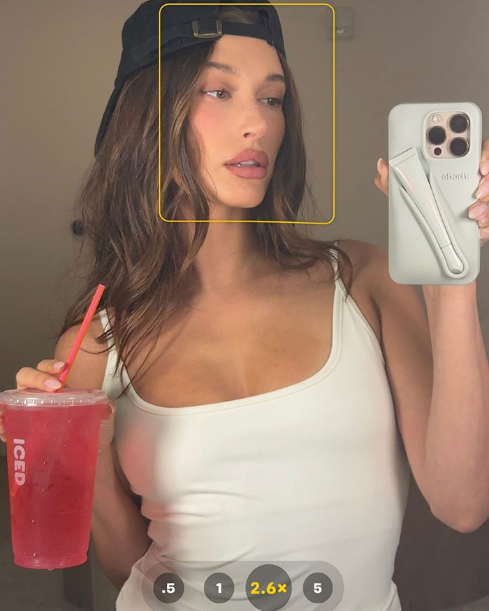 Hailey Bieber tomando una selfie de bikini con una tapa blanca y una gorra negra, sosteniendo una bebida helada roja con una pajita.