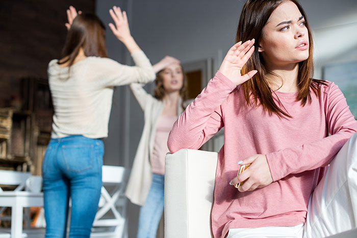La mujer con camisa rosa se ve molesta, mientras que dos hijas discuten en el fondo, ilustrando el conflicto familiar que involucra a medio hermano.