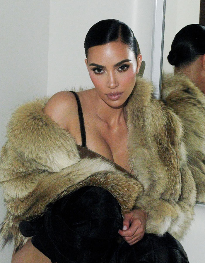 Kim Kardashian con un abrigo de piel posando frente a un espejo con discusiones sobre cirugías de plástico a su alrededor.