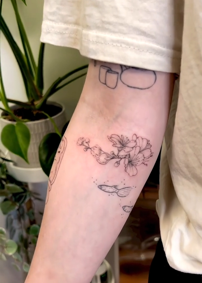 Artista del tatuaje dibujando delicados tatuajes de flores y pescado en el antebrazo de una persona en medio de plantas de interior, reflejando tendencias de tatuaje de tatuaje.
