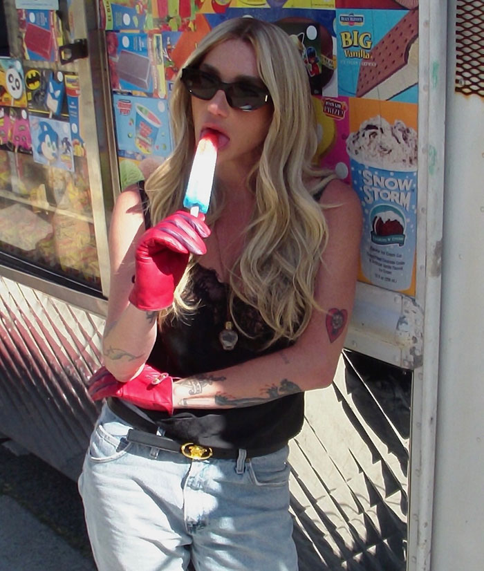 Kesha en gafas de sol y guantes rojos disfrutando de una paleta, en medio de coloridos anuncios y carteles de camiones de helados.