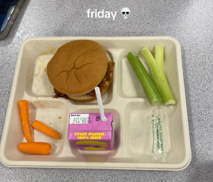 Bandeja de almuerzo escolar estadounidense con un sándwich, zanahorias para bebés, palitos de apio y una caja de jugo de fruta en una mesa.