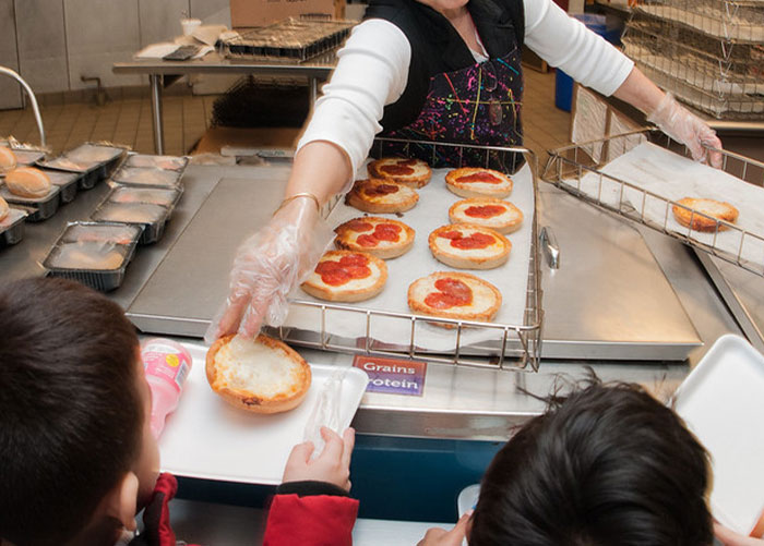 El personal del almuerzo escolar que sirve almuerzos escolares estadounidenses con pizzas mini pepperoni para niños en una línea de cafetería.