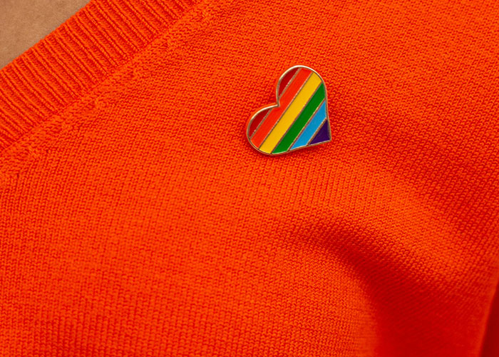 Pin de arco iris en forma de corazón en un suéter naranja, simbolizando la franqueza y las emociones alrededor del viaje con un conflicto de amigos.