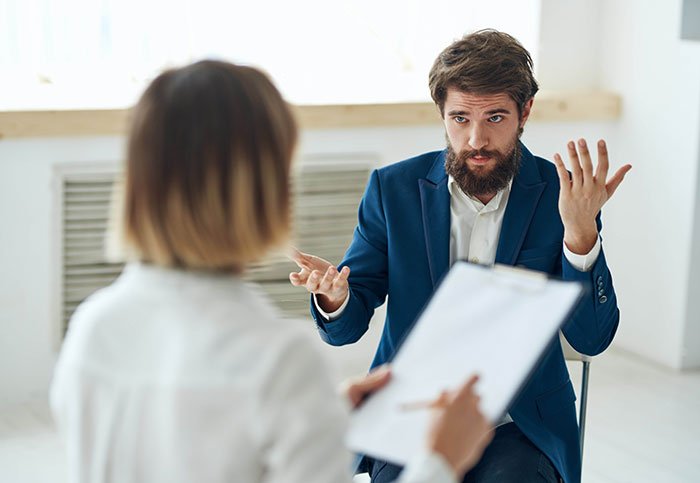 Man in Blue Suit explicando mientras que la mujer con portapapeles escucha en un lugar de trabajo, discusión de disputas para la hora del empleado sin fines de lucro.