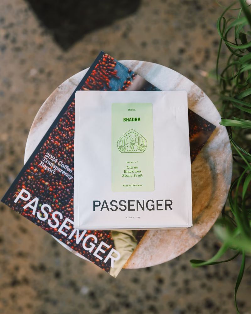 El Insider 23/05/2025: un taburete de madera está cubierto con el informe de transparencia 2024 de Passenger Coffee, un folleto similar a una revista, junto con una bolsa de café de pasajeros etiquetado con notas de degustación de cítricos, té negro y fruta de piedra.