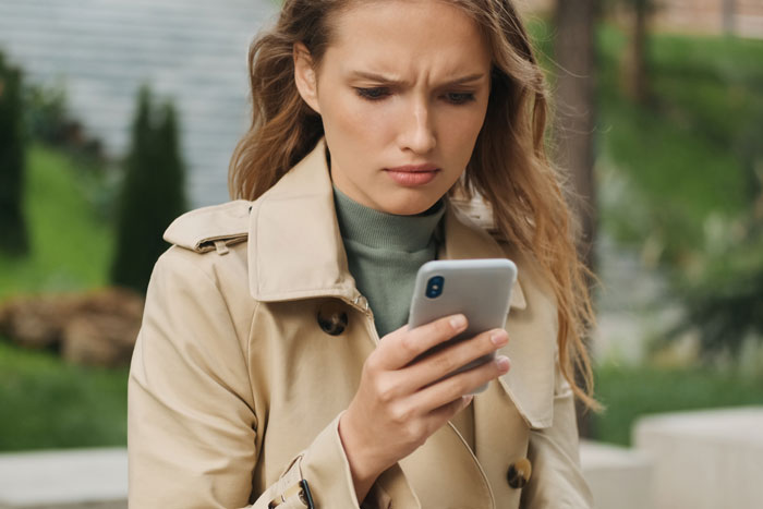 Mujer joven con abrigo beige que parece molesto mientras revisa su teléfono, relacionado con negarse a pagar el problema de la bizca de gato.