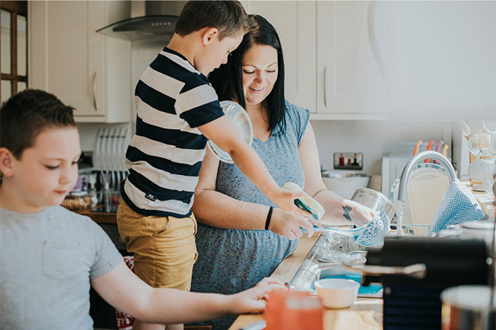 Mujeres y niños que lavan los platos juntos en la cocina, destacando el esfuerzo familiar en medio de desafíos desordenados del hogar.