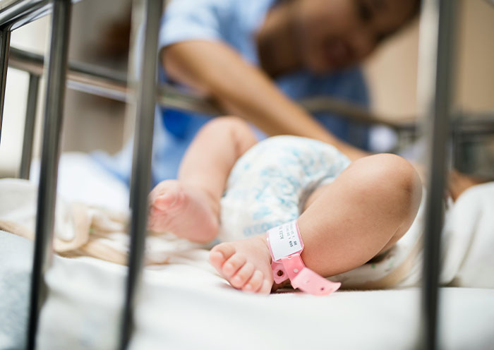 Nueva madre con bebé en la cuna de la UCIN, centrándose en las piernas del bebé con la pulsera del hospital, destacando una situación de vecino de UCIN.