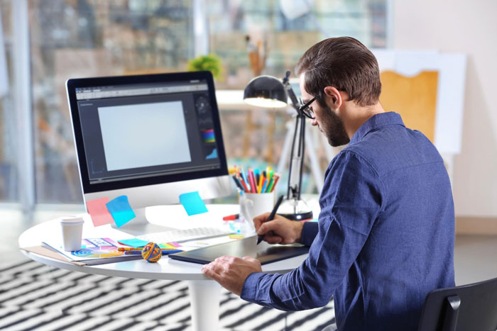 Diseñador que trabaja en tableta digital en el escritorio con computadora, centrándose en las aprobaciones de los clientes y los seguimientos de diseño.