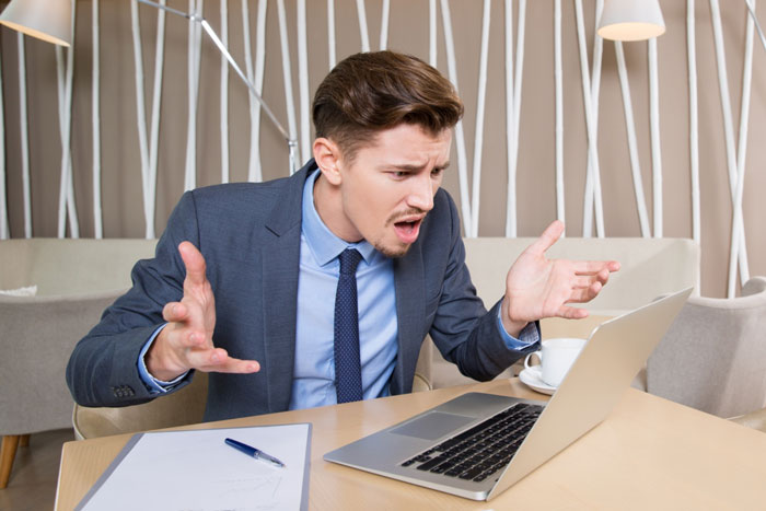 El cliente frustrado reaccionando a la falta de seguimientos de diseñadores en las aprobaciones según lo solicitado, sentado en un escritorio con computadora portátil.