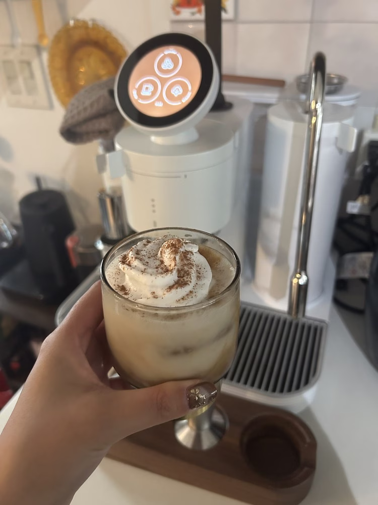 Un primer plano de alguien que sostiene un vaso, que está lleno de hielo, café expreso y crema batida, frente a la máquina de café espresso de Meraki en blanco.