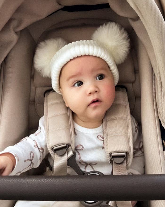 Bebé de 5 meses sentado en un cochecito con un sombrero blanco con pompones, atado de forma segura y mirando a un lado