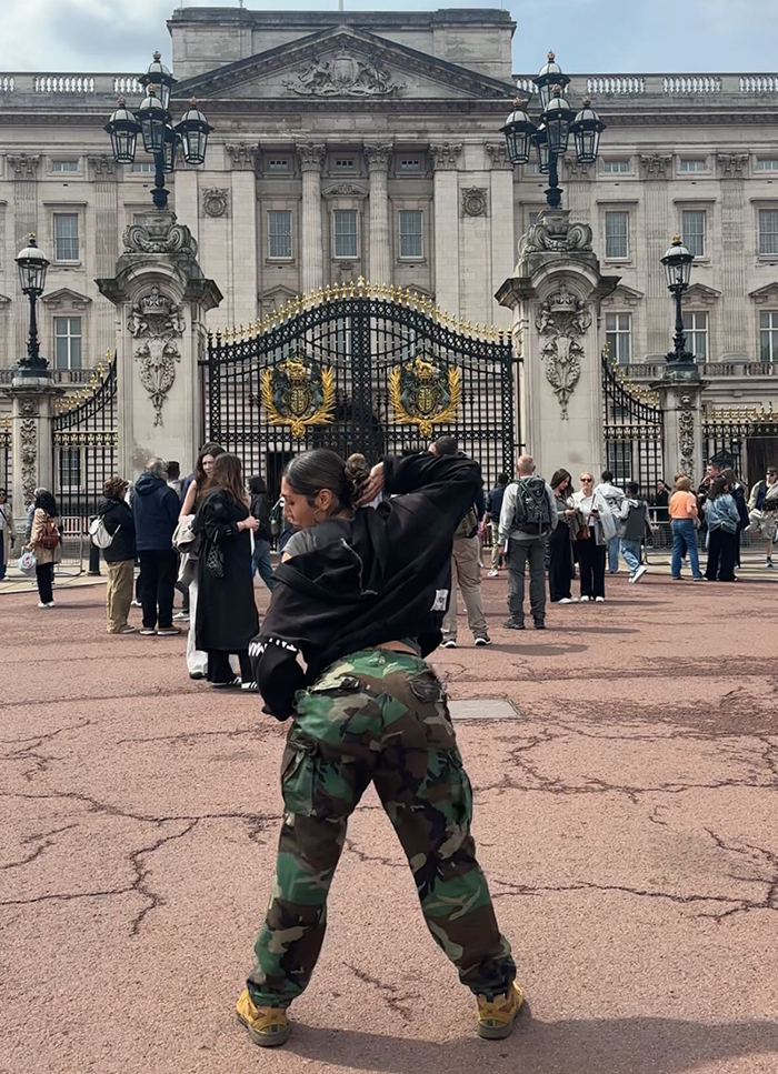 Persona en pantalones de camuflaje posando frente a las puertas del Palacio de Buckingham, relacionado con el artista aborigen y el debate del rey Charles.
