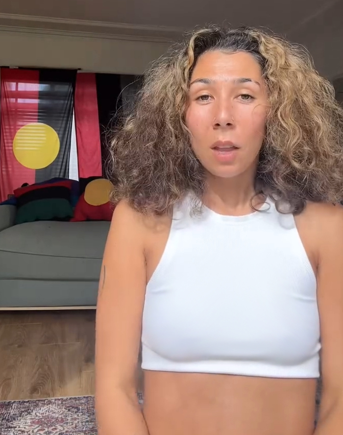 Artista aborigen con cabello rizado en top blanco, hablando en interiores con bandera aborigen y cojines en el fondo.