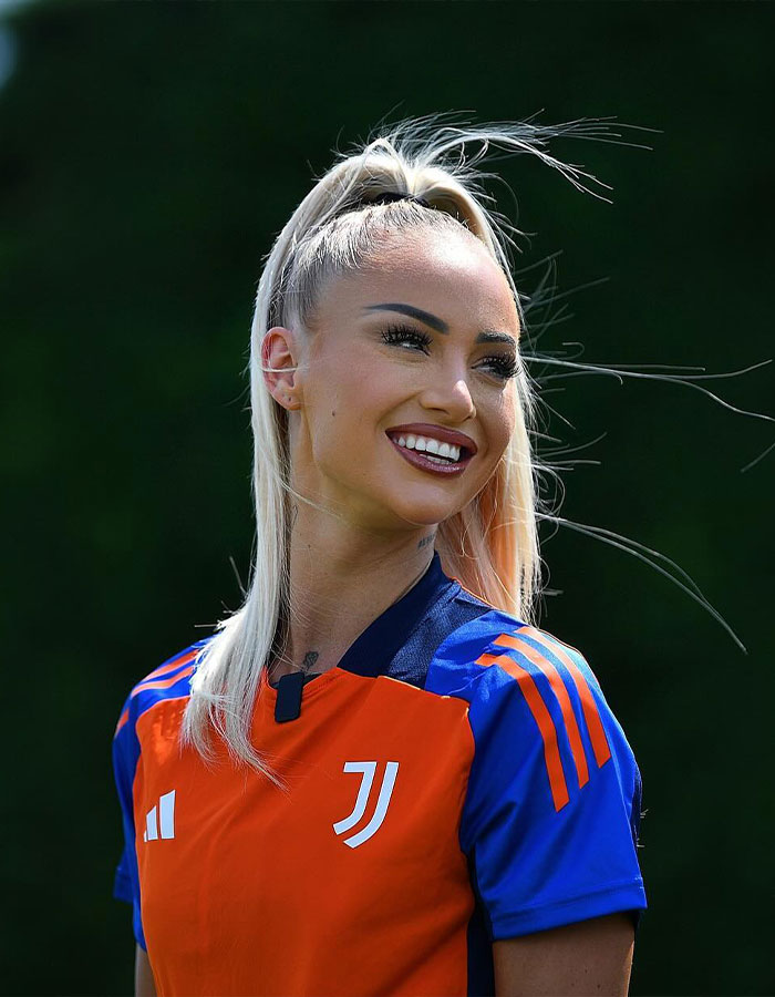 Jugador de equipo nacional de fútbol femenino sonriendo durante la sesión de entrenamiento al aire libre, con camiseta deportiva naranja y azul.