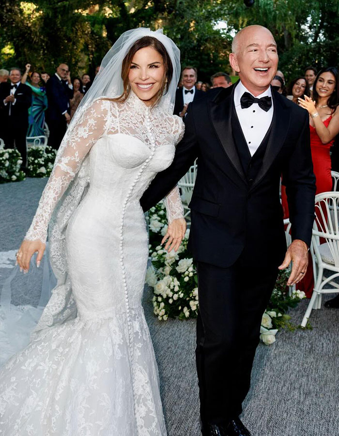 Novia con un vestido de novia de encaje blanco y novio en un esmoquin negro sonriendo mientras camina por el pasillo en una celebración de la boda.