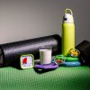 Una vida muerta fotográfica de los objetos utilizados para la recuperación y el enfriamiento posterior al entrenamiento, incluida una gran botella de agua de metal amarillo, un rodillo de espuma negra, un bloque de yoga negro, un pequeño plato de vidrio de almendras, un pequeño plato de vidrio de galletas de pez dorado, un vaso de leche, un temporizador visual de color verde lima a 20 minutos y unos pocos placos de pesas, todos arreglados en un paso de ejercicio cubierto con una matera de caña de cótlo verde con una matera de caña verde.