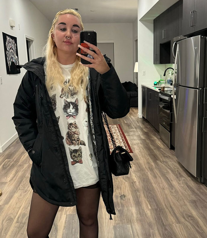 Amanda Bynes tomando una selfie de espejo en el interior, con una chaqueta negra y una camisa gráfica de gato, relacionada con las noticias de uso de Ozempic.