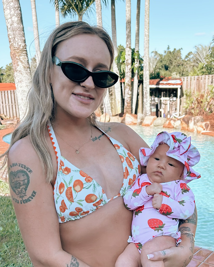 Mujer con gafas de sol sosteniendo un bebé de 5 meses en trajes de baño de fresa junto a una piscina con palmeras en el fondo.