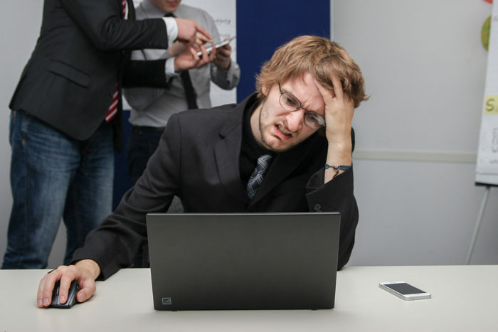 El hombre frustrado en gafas lucha en la computadora portátil, mientras que los colegas discuten en segundo plano, ilustrando el pánico del cliente después de la aprobación de la aprobación de seguimiento.