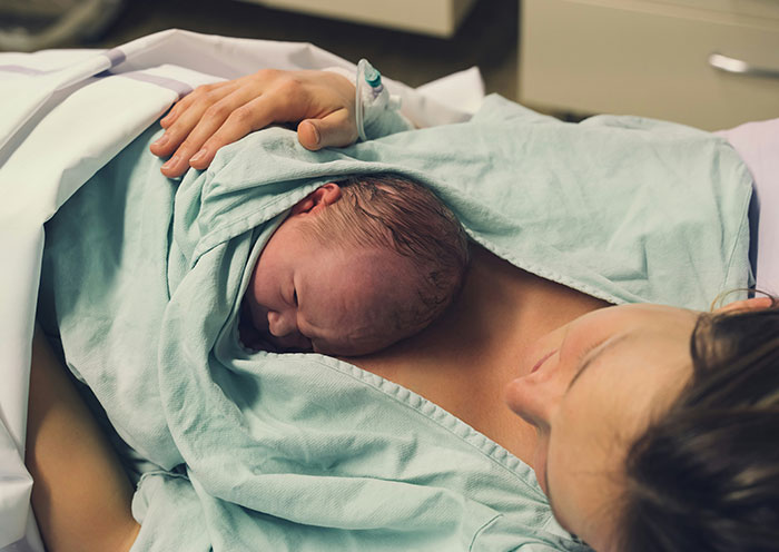 Nueva madre que sostiene al recién nacido en la cama del hospital, ilustrando una experiencia de la UCIN con un vecino entrometido y un cumplimiento malicioso.