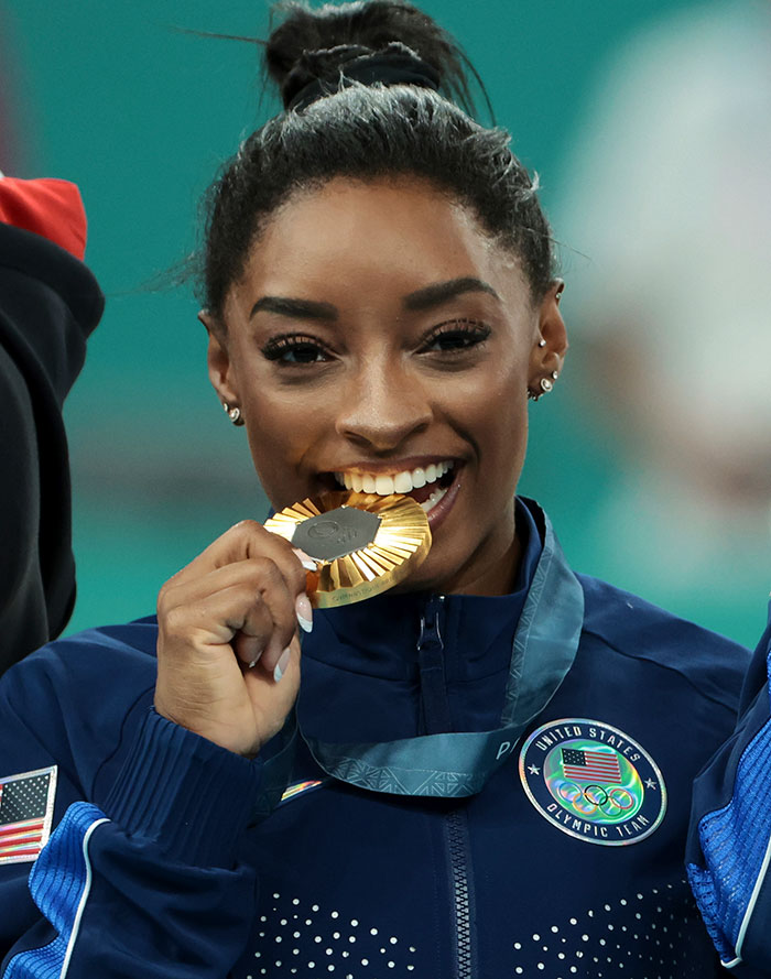 Simone Biles sonriendo y mordiendo una medalla de oro mientras usa la chaqueta del equipo de EE. UU., Provocando rumores de cirugía plástica