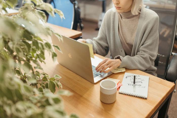 Mujer que trabaja en una computadora portátil en un escritorio, enfocada en escribir, con cuaderno y taza de café cerca en un acogedor espacio de trabajo.