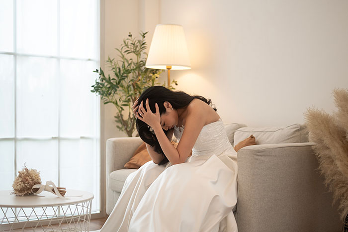 Novia con un vestido blanco sentado en un sofá de tonos neutros que se ve molesto en una sala de estar suavemente iluminada.