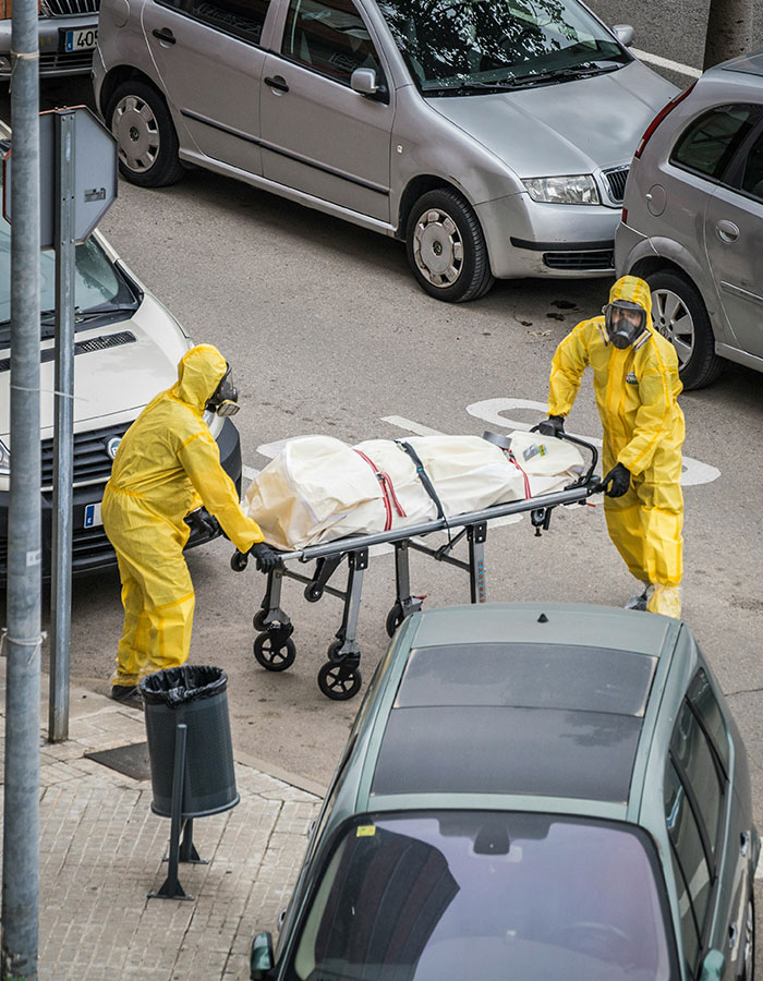 Dos trabajadores con trajes de peligro que transportan una bolsa de cuerpo en una camilla que destaca las formas inusuales en que las personas han fallecido.