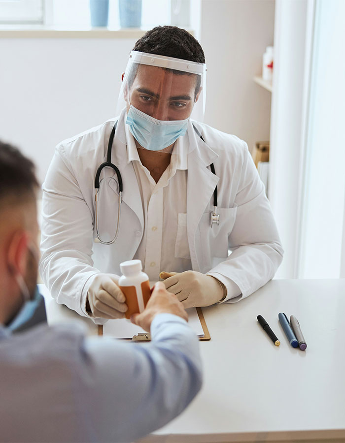 Profesional médico que usa una máscara y un escudo facial que entrega una botella de medicamentos a un paciente en un entorno clínico relacionado con la tecnología de la autopsia.