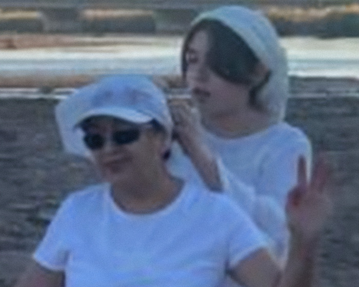 Dos mujeres al aire libre cerca del agua, una arreglando el pañuelo en la cabeza del otro, con expresiones tranquilas en un entorno informal.
