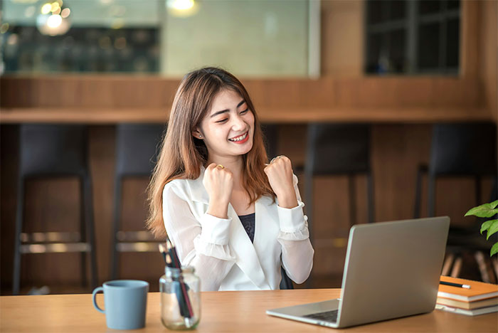 Mujer joven con doctorado celebrando el éxito en la computadora portátil en el espacio de trabajo de interior moderno con taza de café y cuadernos en la mesa