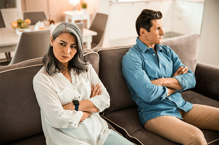 La mujer con doctorado se ve molesta sentada frente a su esposo en el sofá con los brazos cruzados en la brillante sala de estar moderna.