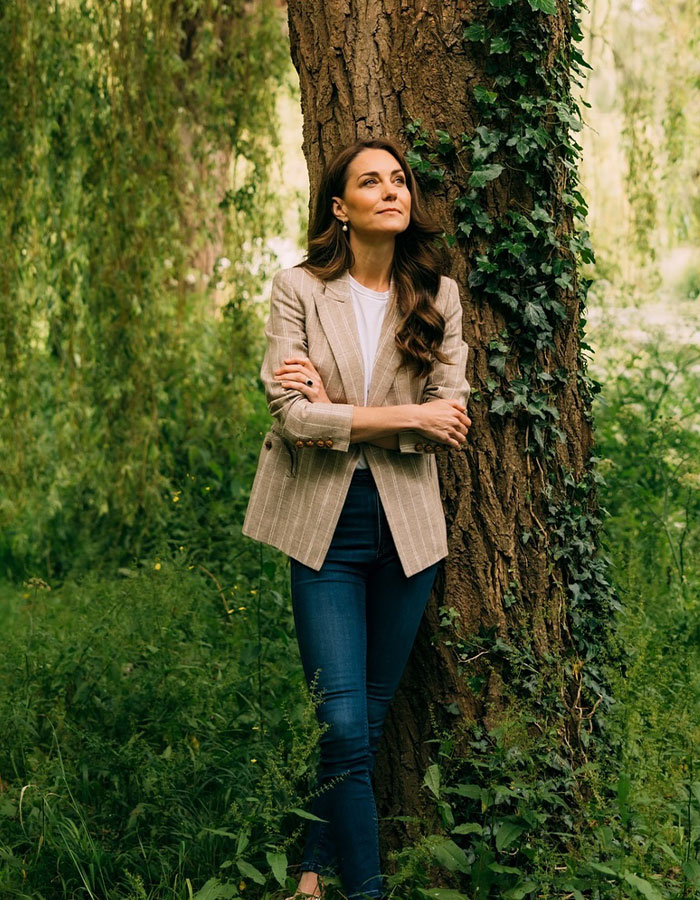 La ex altura asistida de Kate Middleton de pie en árbol en blazer casual y jeans, luciendo reflexivo en un entorno verde al aire libre.