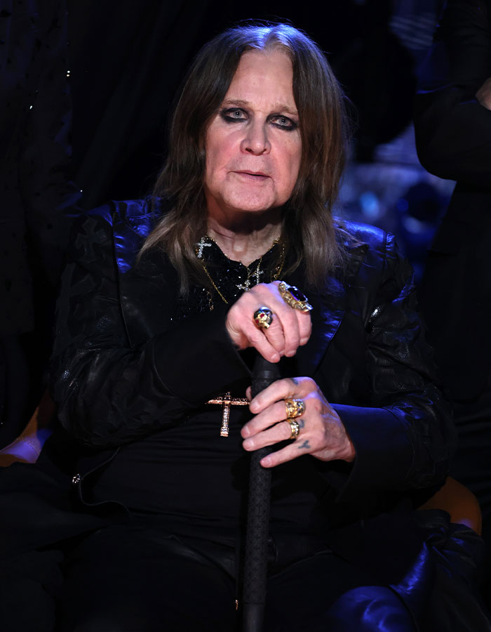 Ozzy Osbourne con cabello largo y maquillaje oscuro, vistiendo anillos y sosteniendo un bastón en un entorno con poca luz.