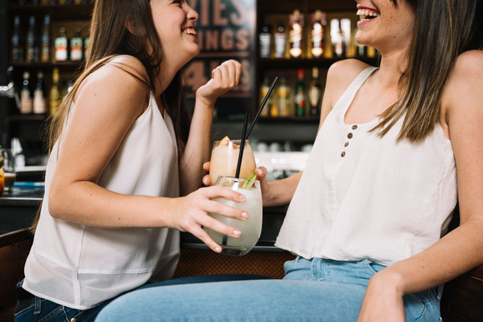 Dos mujeres se ríen y sostienen cócteles en un bar, ilustrando una escena de una prometida que bebe y un incidente de lesiones.
