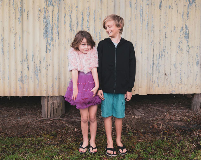 Dos hermanos de pie afuera, el niño dorado con vestido morado que se ve tímido mientras el hermano la sonríe.