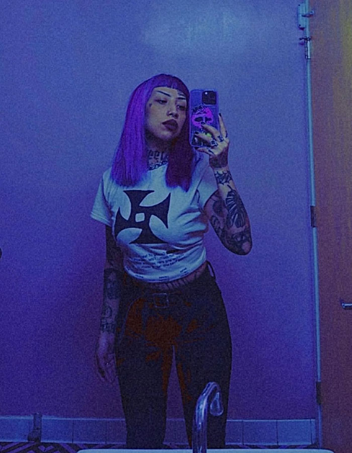 Mujer joven con cabello morado y tatuajes tomando una selfie espejo en una habitación con poca luz, simbolizando preocupaciones de planificación de disparos escolares