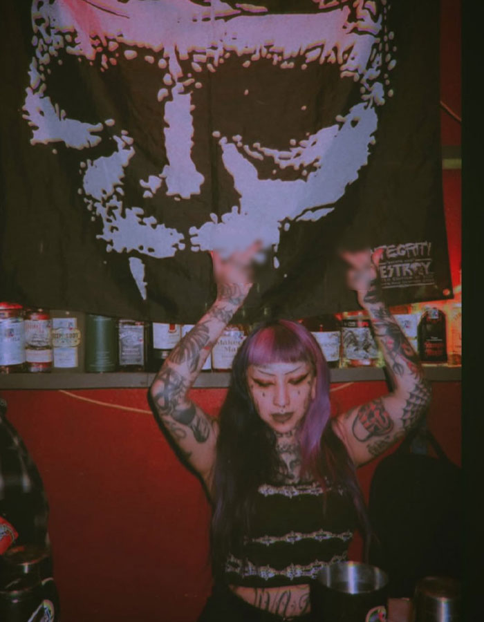 Persona con tatuajes y cabello púrpura levantando las manos en una habitación con poca luz, que ilustra los detalles repugnantes sobre los planes de tiro en la escuela.