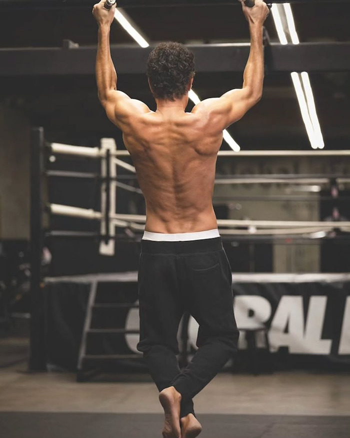 El hombre musculoso realizando pull-ups en un gimnasio, destacando un físico del cuerpo Marvel relacionado con la controversia de Sacha Baron Cohen.
