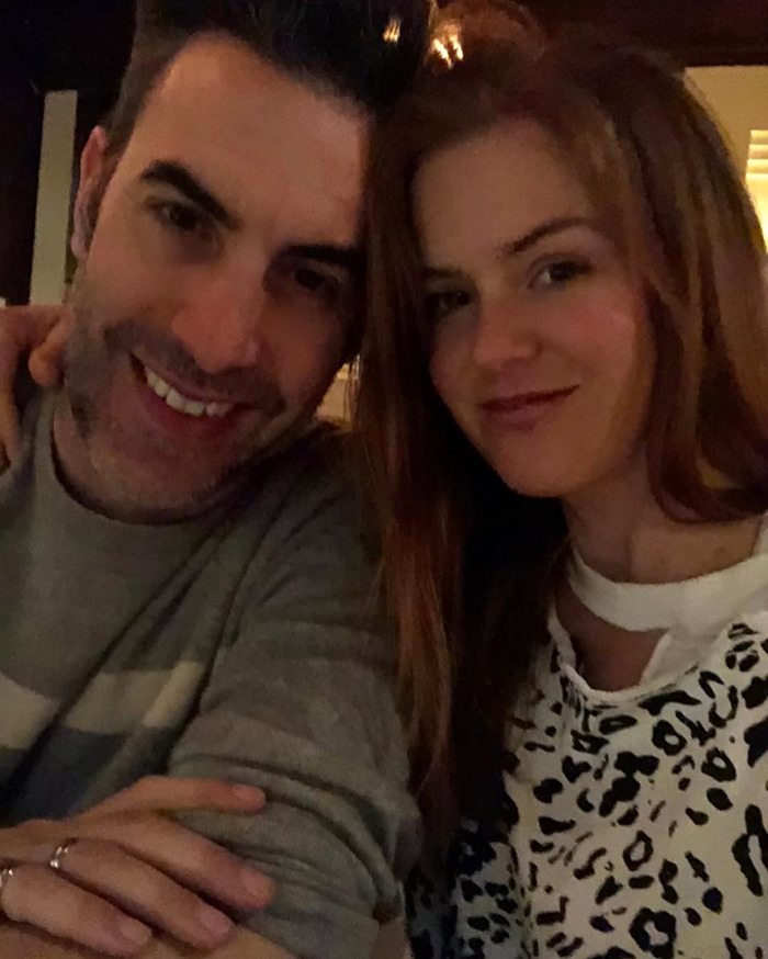 Hombre y mujer sonriendo juntos en un entorno con poca luz, relacionados con la reacción de columnista al cuerpo de Sacha Baron Cohen Marvel.