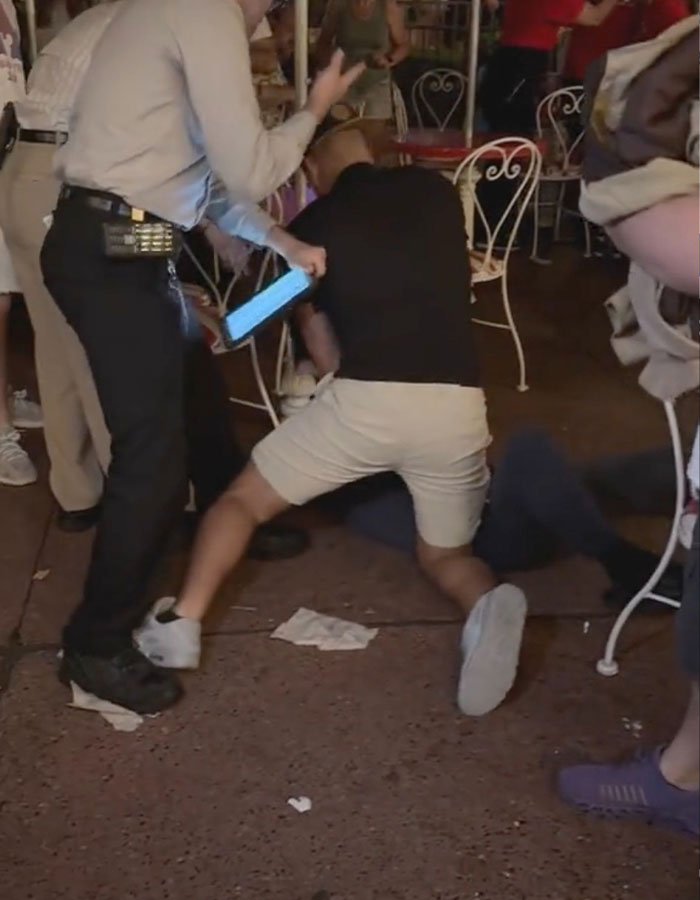El hombre con camisa negra agachada sobre otra persona durante un violento incidente del mundo de Disney con espectadores cercanos.