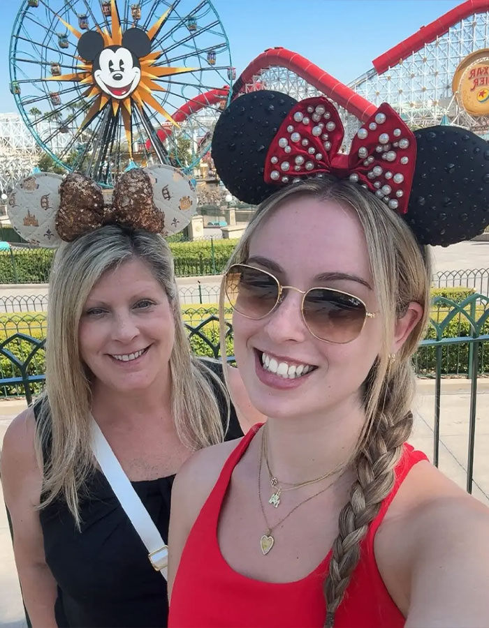 Dos mujeres con orejas Minnie Mouse en Disney World con una rueda de la fortuna y una montaña rusa en el fondo durante el día.